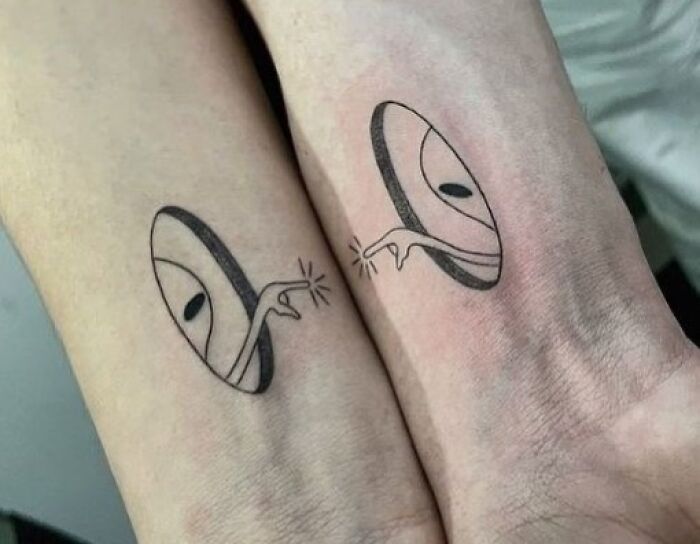 two people with matching tattoos on their arms, one has an alien face and the other has