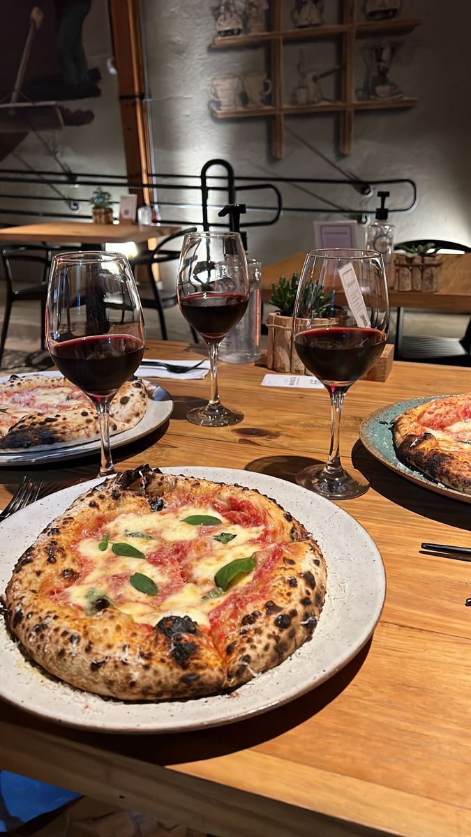 two pizzas sitting on top of a wooden table next to glasses of red wine