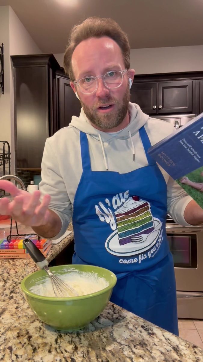 a man in an apron holding a cookbook and whisk next to a bowl of food