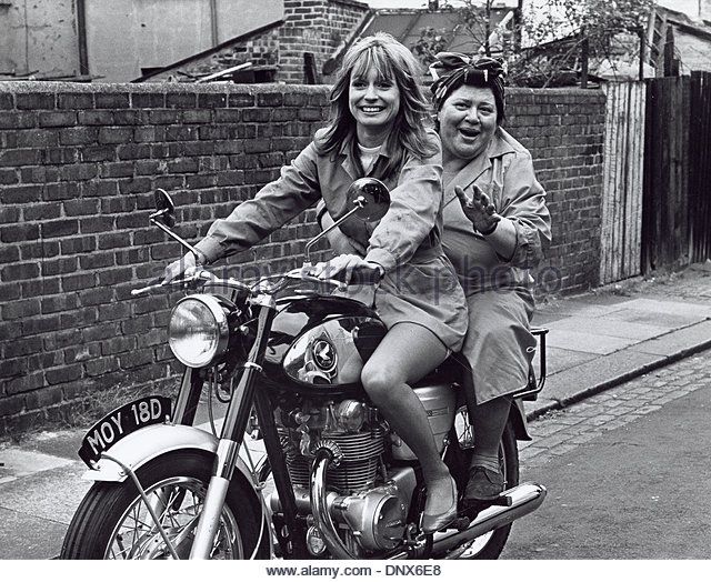 two women riding on the back of a motorcycle in front of a brick wall and building