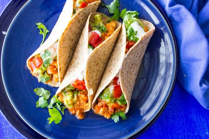 three tacos on a blue plate with cilantro, tomatoes and lettuce