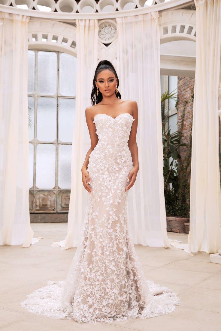 a woman standing in front of a window wearing a wedding dress with flowers on it