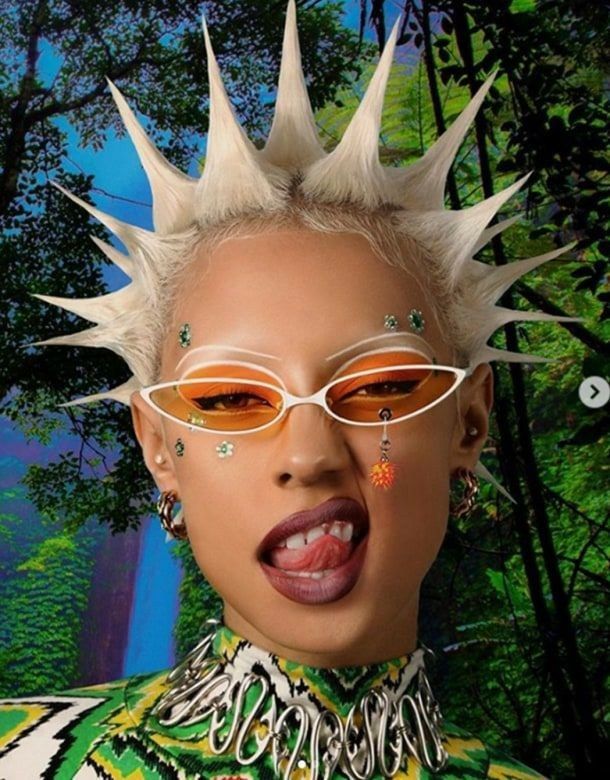 a woman with spiked hair and glasses in front of a forest background, wearing an unusual outfit