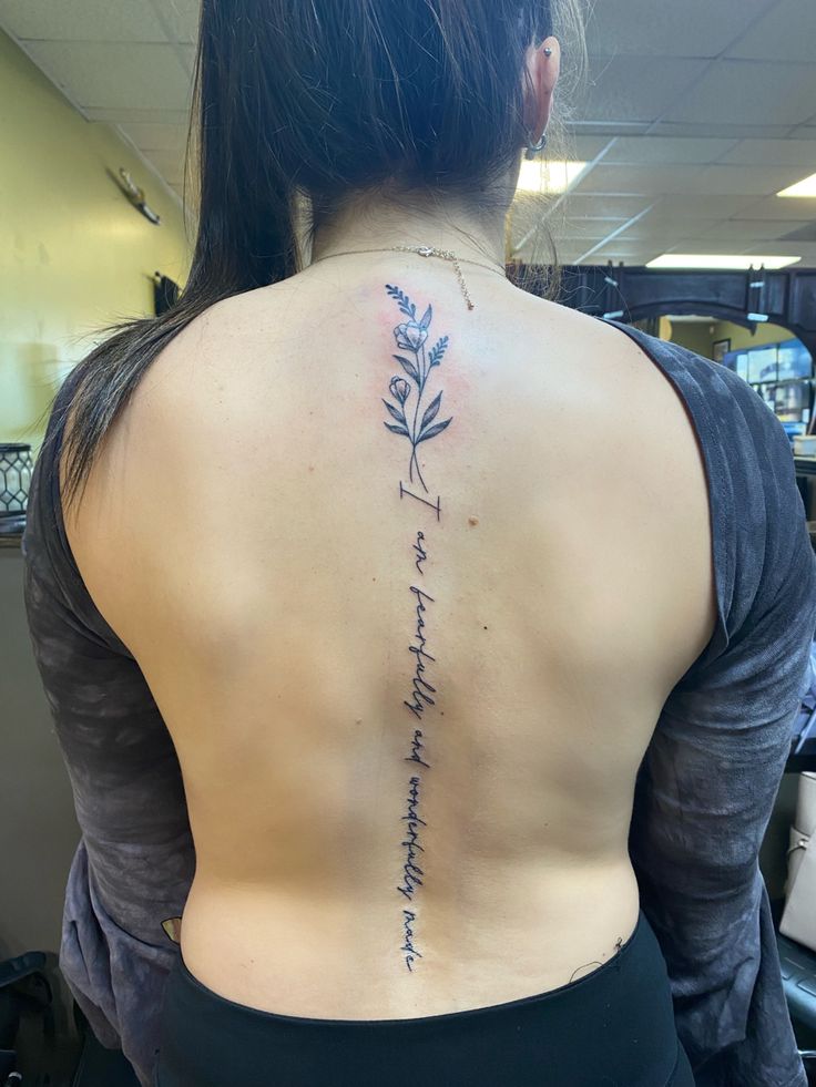 the back of a woman's body with a tattoo on her left shoulder and writing in cursive font