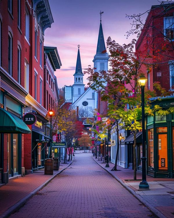 a city street lined with tall red brick buildings at dusk, with the steeple of a church in the background