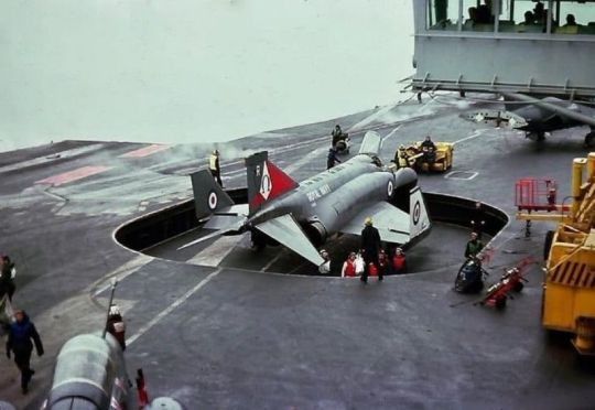 an airplane on the deck of a ship with people standing around and looking at it