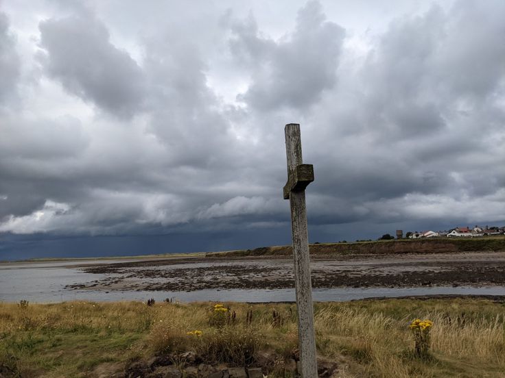 a wooden cross in the middle of a grassy area with water and clouds behind it