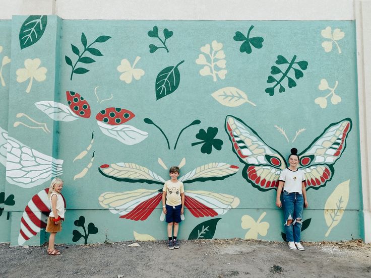 three people standing in front of a painted wall with butterflies and ladybugs on it