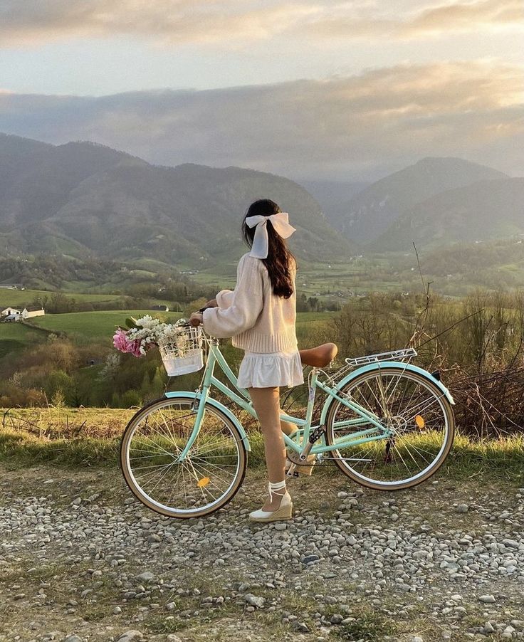 a woman standing next to a blue bike on top of a field with mountains in the background