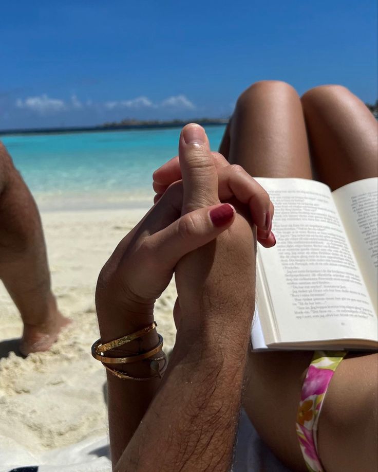 a person laying on the beach with a book in their hand and two other people behind them