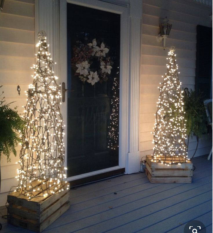 two christmas trees are on the porch with lights around them and in front of a door