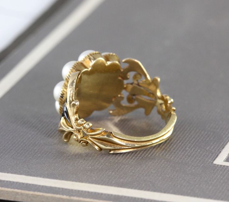 "An immaculate antique Georgian mourning ring, crafted in 18k yellow gold with a lustrous split pearl halo around the center compartment which holds a momento of woven hair under crystal. The shoulders are delicately articulated in a baroque scroll design, accented by glossy black enamel, and the ring is fully hallmarked for 18k, London manufacture, and letter dated 1828. Measuring approximately 1/2\" north to south at the center front, tapering through the shoulders to 2.22 mm at the center bac Antique Yellow Gold Pearl Ring, Antique Yellow Gold Pearl Ring With Rose Cut Diamonds, Victorian Yellow Gold Pearl Ring With Rose Cut Diamonds, Antique Cameo Signet Ring For Weddings, Victorian 14k Yellow Gold Pearl Ring, Victorian Style 14k Yellow Gold Pearl Ring, Victorian Enamel Ring For Anniversary, Antique Style Round Enamel Wedding Ring, Antique Yellow Gold Enamel Wedding Ring