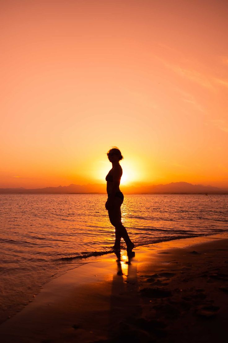 the silhouette of a woman standing on top of a beach next to the ocean at sunset