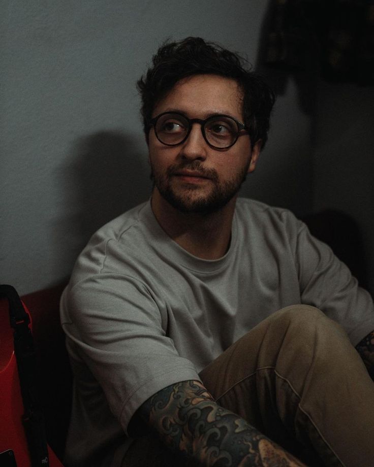 a man with tattoos and glasses sitting in a chair looking at the camera while wearing eyeglasses