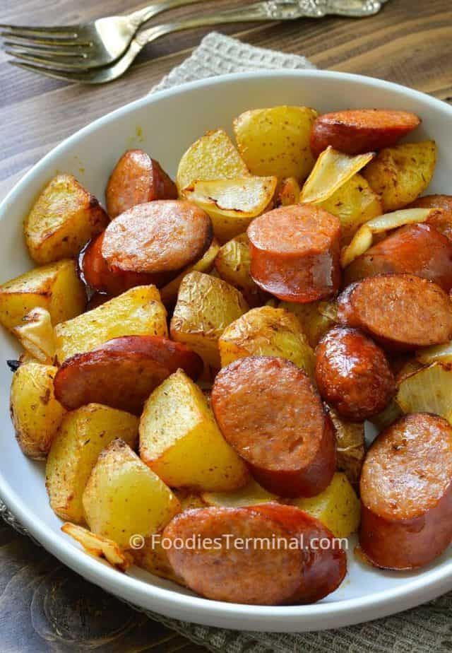 a white bowl filled with potatoes and sausages on top of a wooden table next to a fork