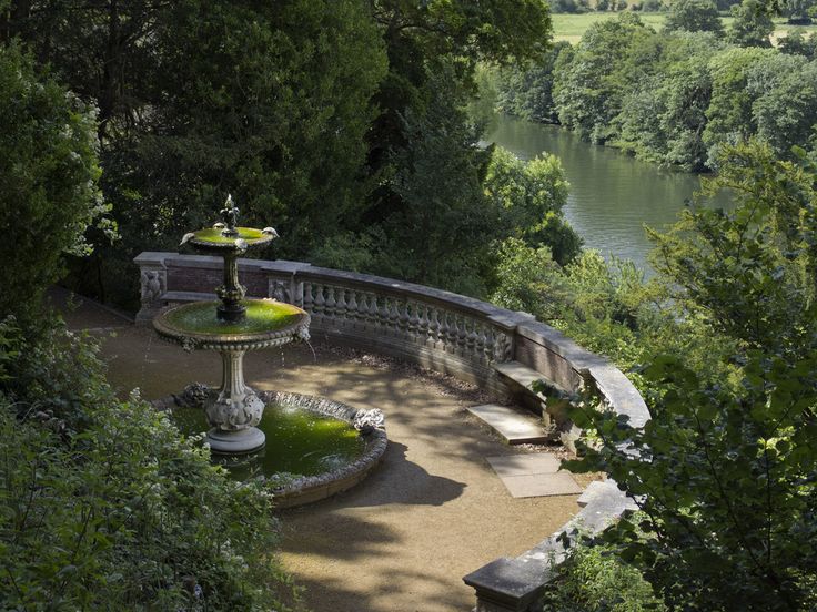 a fountain in the middle of a garden with trees around it and water behind it