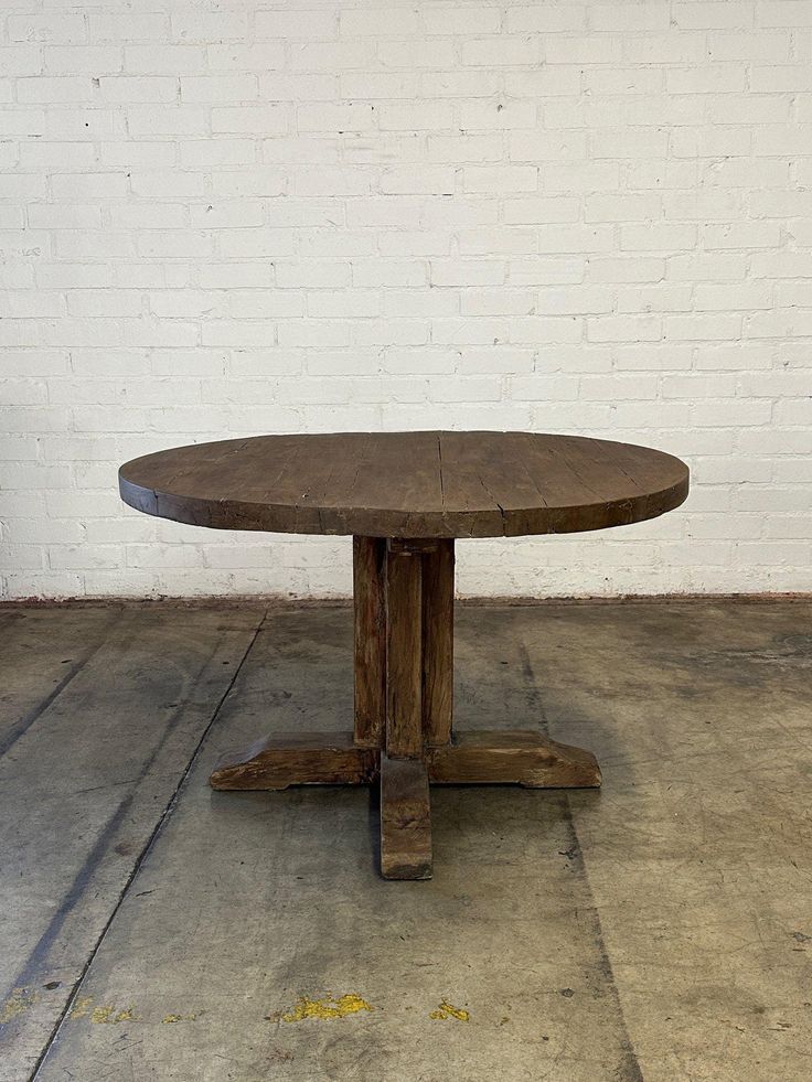 a round wooden table sitting on top of a cement floor next to a white brick wall