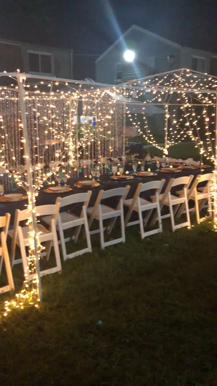 rows of white folding chairs with fairy lights on them at an outdoor wedding reception in the evening