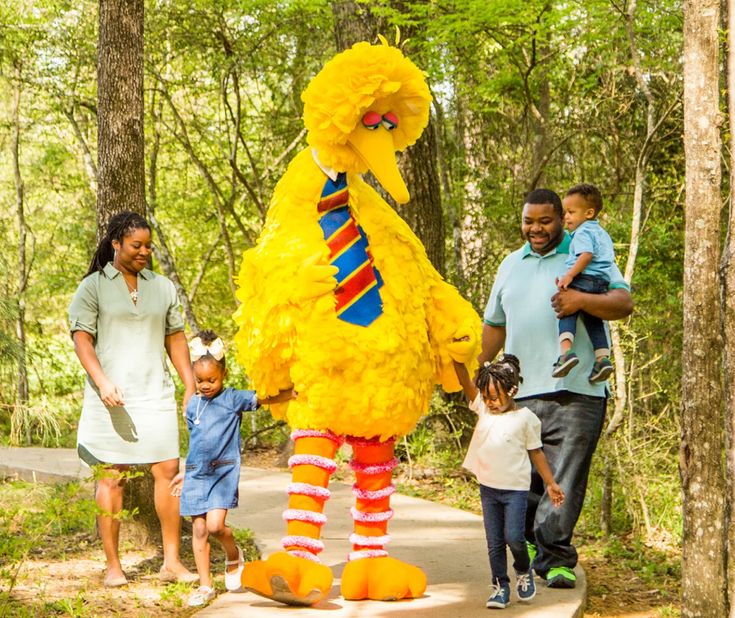 a group of people standing next to a large yellow bird in the middle of a forest