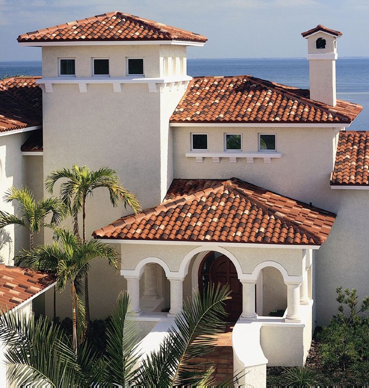 a large white building with red tile roofing next to the ocean and palm trees