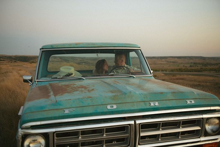 two people sitting in the front seat of an old pickup truck on a dirt road