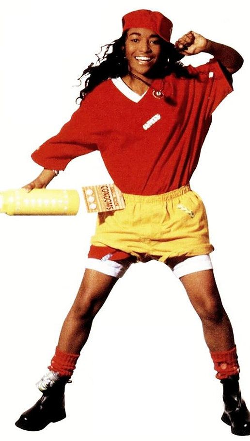 a woman in red shirt and yellow shorts holding a baseball bat with both hands on her hip