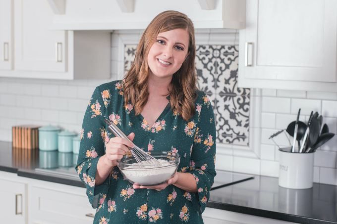 a woman standing in a kitchen holding a bowl with whisk and spoons