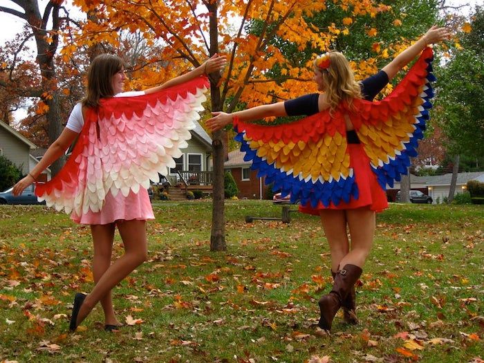 two young women dressed in colorful paper wings dancing on the grass with trees and leaves behind them