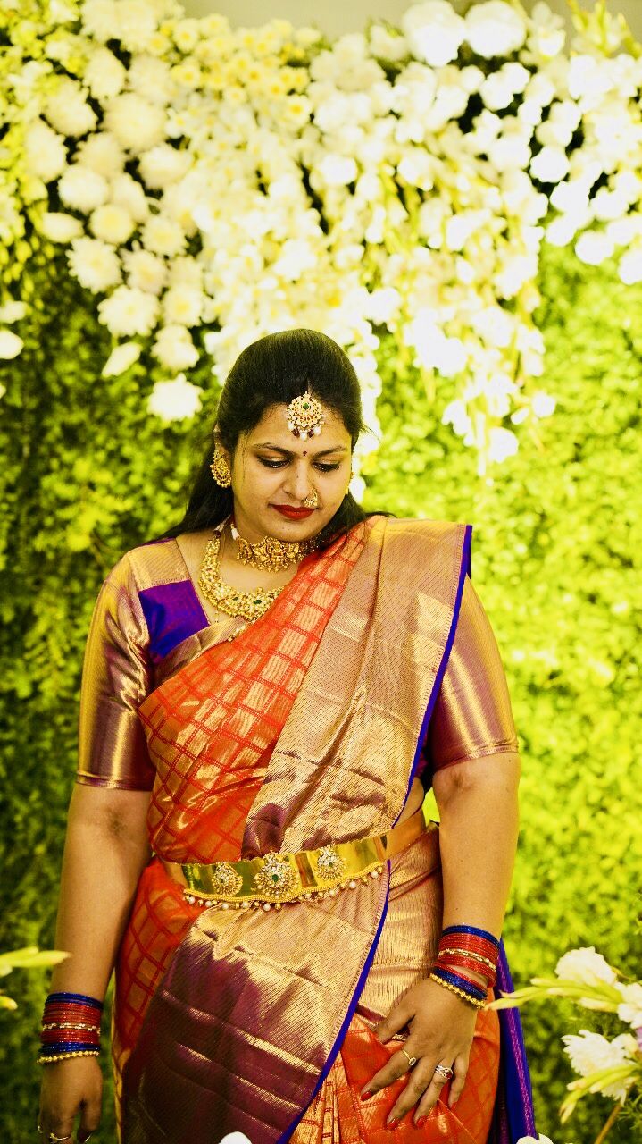 a woman in an orange and purple saree standing next to flowers with her hands on her hips