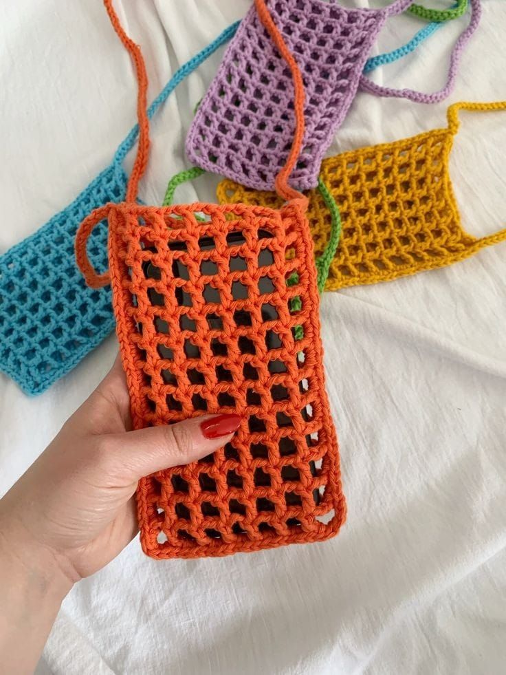 a hand holding an orange crocheted purse on top of a white bed next to four different colored bags