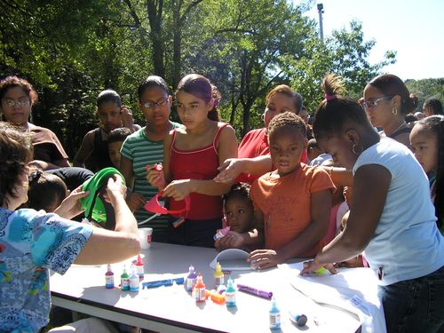 a group of people standing around a white table with bottles on it and one person holding something in the other hand