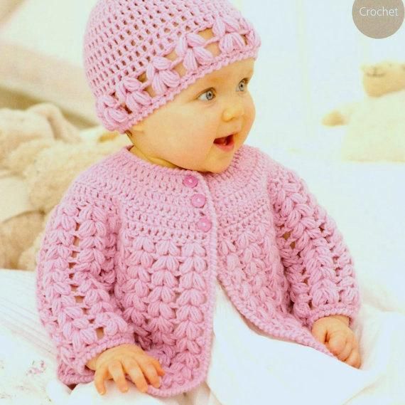 a baby wearing a pink crochet sweater and hat sitting on a bed with white sheets