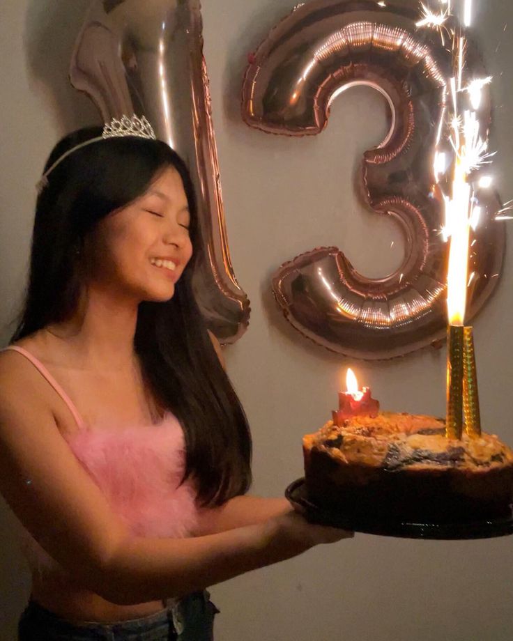 a woman holding a cake with lit candles in front of a number three balloon wall