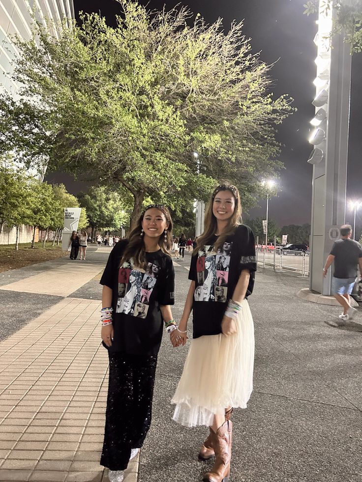 two young women standing next to each other on a sidewalk at night with trees in the background