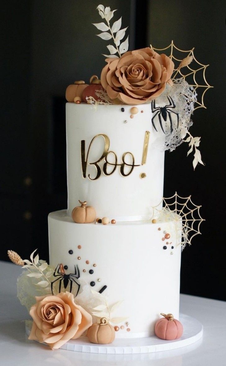 a three tiered white cake with gold lettering and flowers on the top, sitting on a table