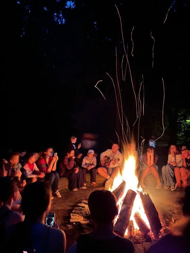 people sitting around a campfire at night while others sit on the ground watching it