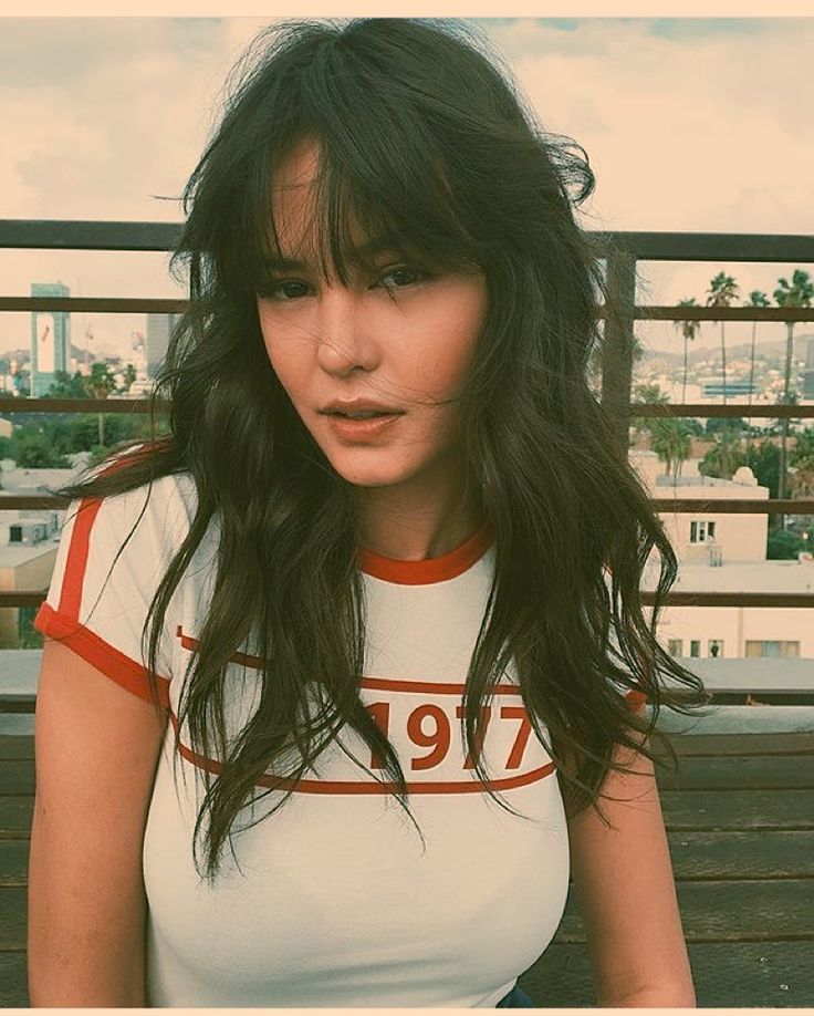 a woman with long black hair wearing a white t - shirt and sitting on a bench