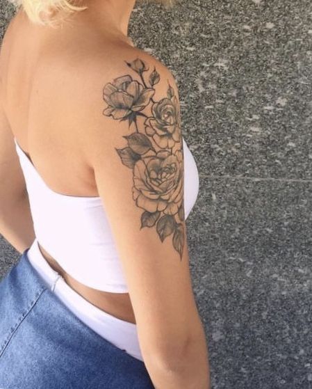 a woman with a flower tattoo on her left arm and shoulder, standing in front of a stone wall