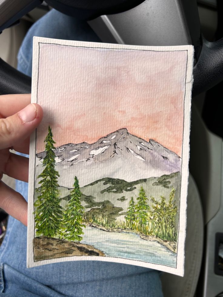 a person holding up a watercolor painting of mountains and trees in front of them