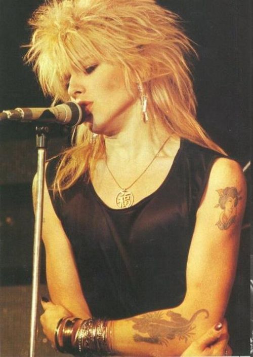 a woman with long blonde hair and tattoos on her arm, singing into a microphone