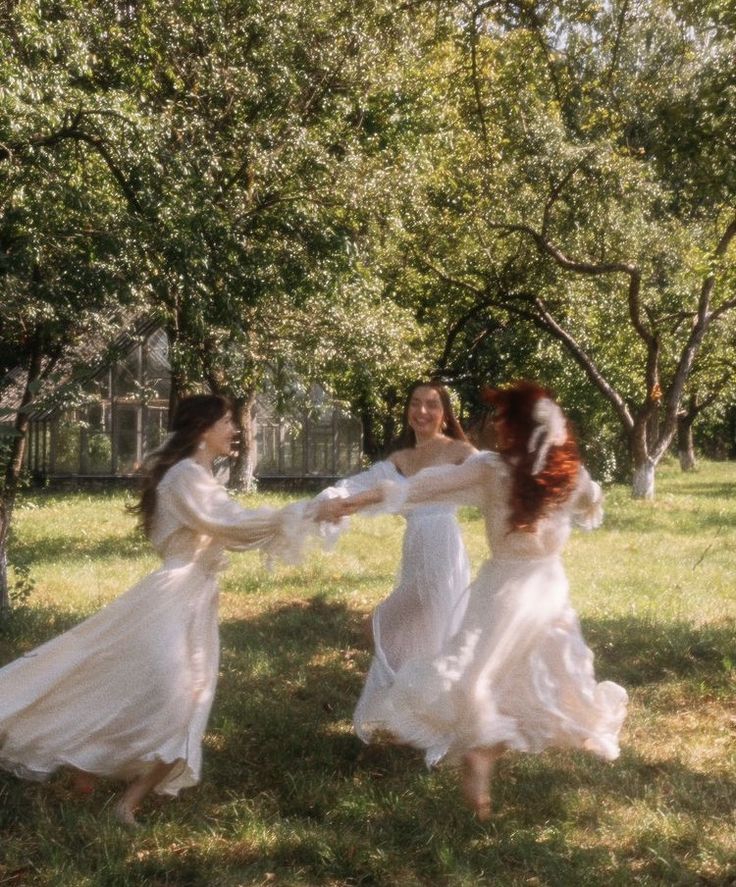 three women in long white dresses are playing with each other on the grass near trees