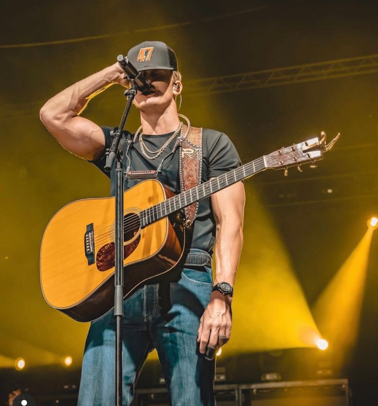 a man holding a guitar on stage with his hands behind his head and wearing a hat