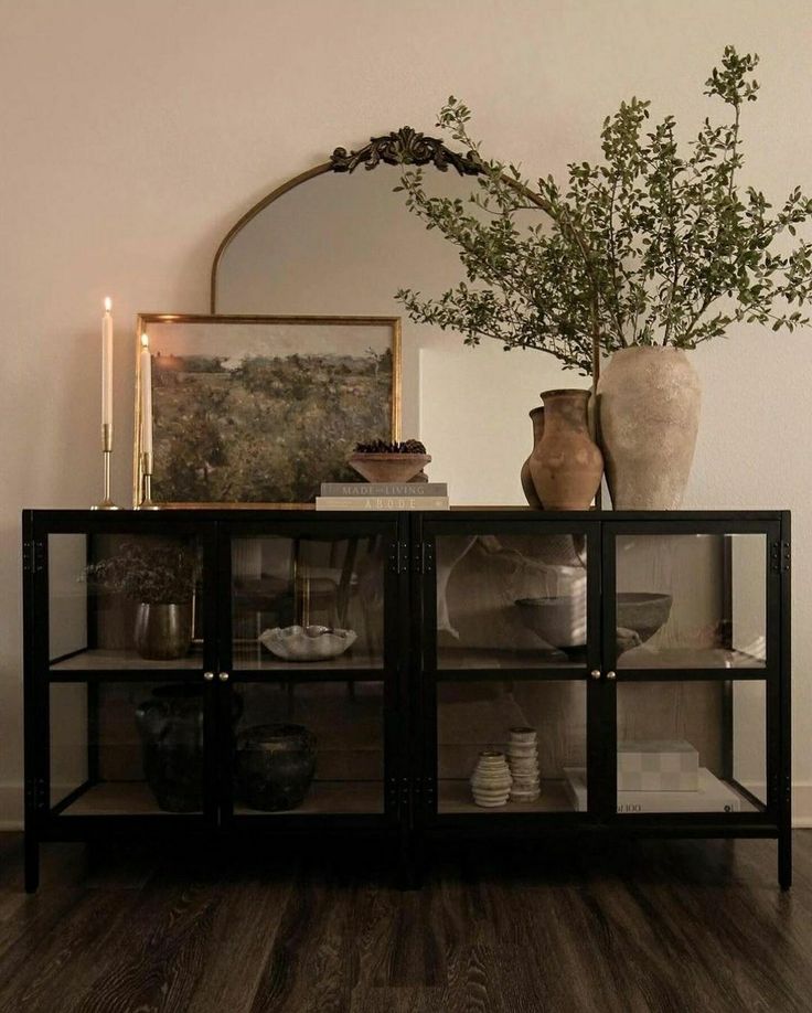 a glass cabinet with vases and plates on it in front of a painting, candles and a mirror