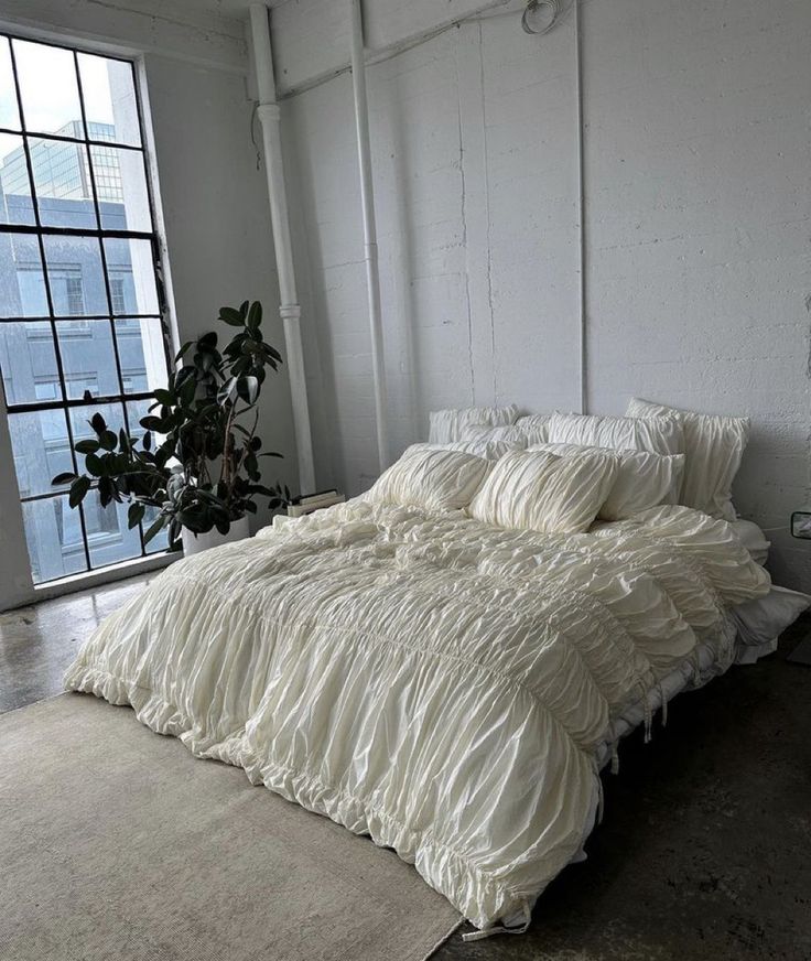 an unmade bed sitting in front of a window next to a potted plant