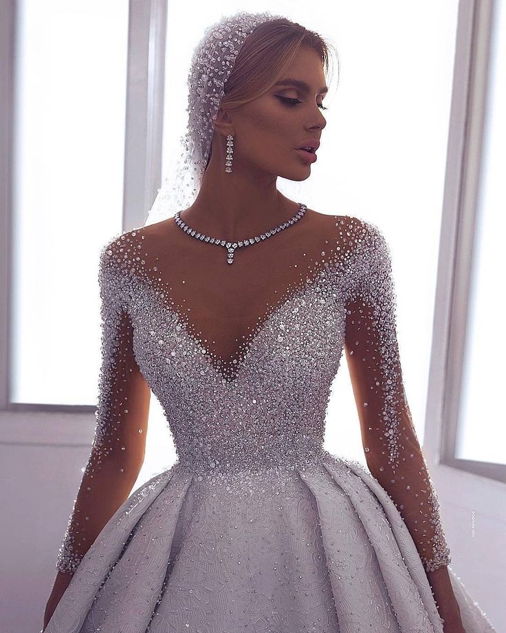 a woman in a wedding dress with pearls on the neck and long sleeves, standing next to a window