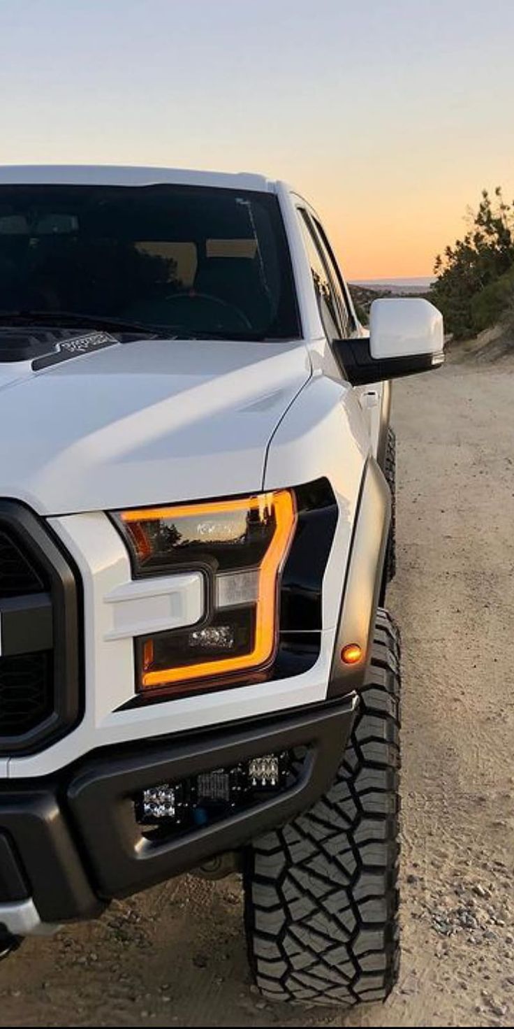 the front end of a white truck parked on top of a dirt road at sunset