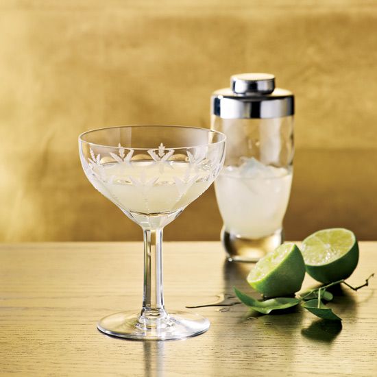 two wine glasses sitting on top of a wooden table next to limes and a shaker