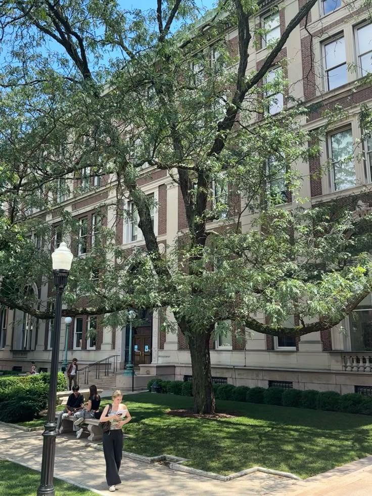 people are sitting on benches in front of a large building with many windows and trees