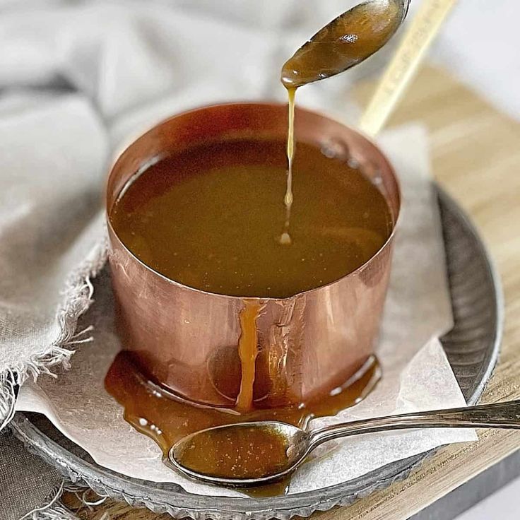 a spoon is pouring caramel sauce into a copper cup on a plate with silverware