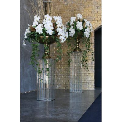 three clear vases with white flowers in them on a gray floor next to a brick wall